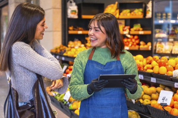 Friendly store employee wearing an apron and gloves holding a digital tablet, helping a smiling