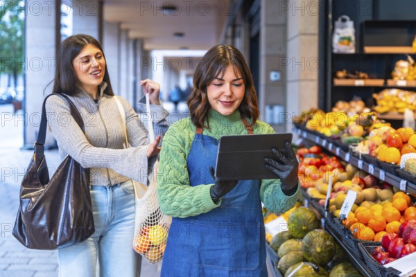 Greengrocer in apron and gloves uses a tablet to manage inventory and assist a smiling customer