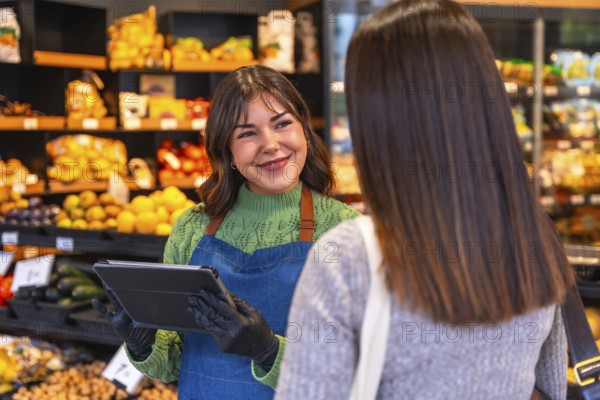 Friendly saleswoman holding a tablet and wearing gloves, assisting a customer with produce in a