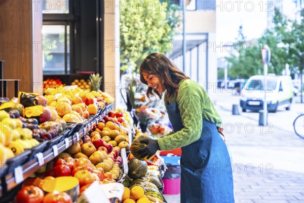 Woman smiling as she arranges colorful fresh fruits and vegetables on an outdoor market stand,
