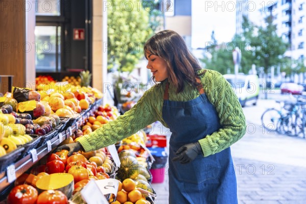 Smiling young greengrocer woman wearing an apron and gloves arranging fresh fruit and vegetables on