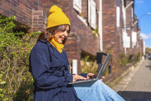 Smiling woman wearing a yellow beanie and blue trench coat, typing on a laptop while sitting