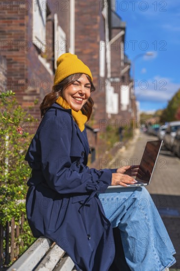 Young woman smiling at camera while sitting on a public bench in an autumn street, typing on her