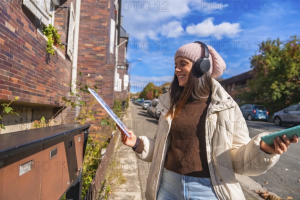 Happy young woman standing on an urban street, opening a mailbox to retrieve mail while wearing