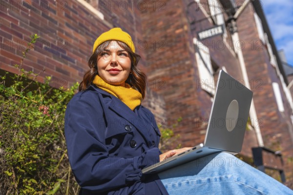 Woman wearing a warm yellow beanie and trench coat, smiling while sitting outdoors with a laptop on