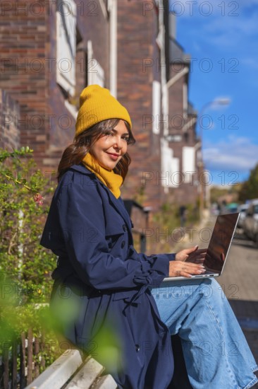 Young woman smiling while sitting on a bench, typing on her laptop, conducting remote work or