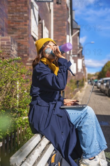 Woman hydrating with a water bottle while working remotely on her laptop, sitting on a bench in an