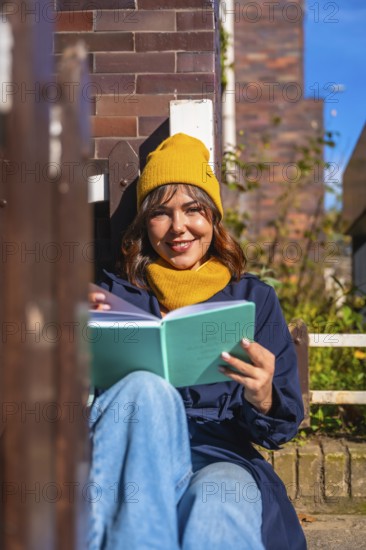 Young woman in a yellow beanie sits on a bench against a brick wall, smiling and reading a book