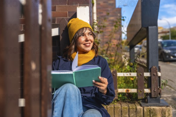 Woman in casual autumn attire reading a notebook while sitting outdoors, smiling and contemplating