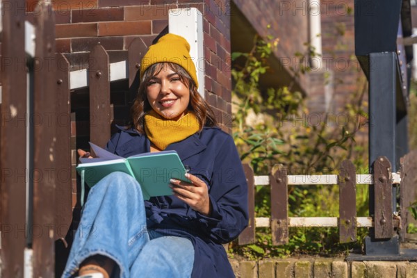 Young woman wearing a yellow beanie and scarf. Sitting outdoors in autumn sunlight. Smiling while