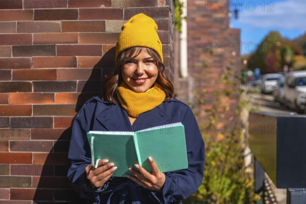 Young woman wearing a yellow beanie and scarf, dressed in a trench coat, standing outdoors in warm