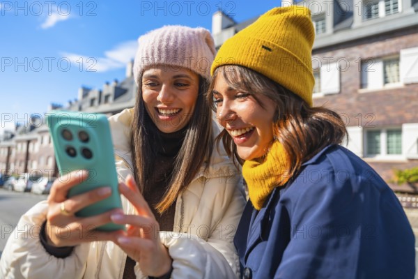 Two young women, dressed in winter hats and warm clothing, are laughing while looking at a mobile