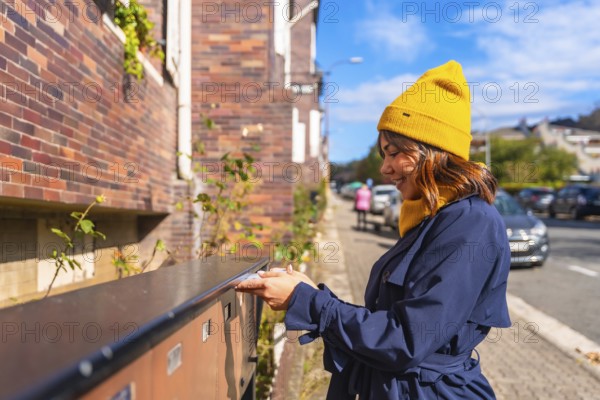 Woman wearing a yellow beanie and blue trench coat, smiling while sending a letter through a street