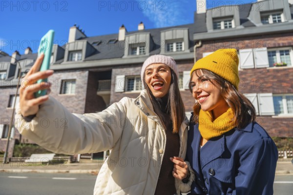 Two cheerful young women friends are enjoying a sunny winter day in the city, smiling and taking a