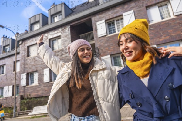 Two young women friends walking a city street in winter coats and beanies, laughing and chatting