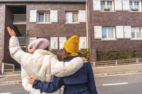 Two young women walking arm in arm down a city sidewalk in cool weather, wearing parkas and