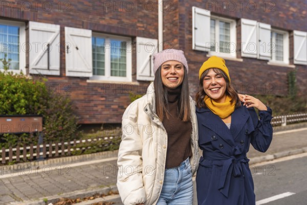 Two smiling young women friends are standing together on an urban street in front of a brick