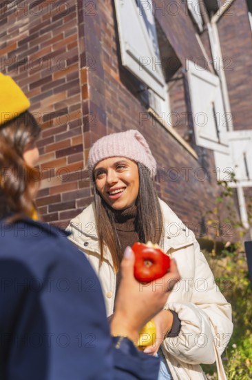 Two young women chatting outdoors on a sunny urban street, smiling and sharing apples while bundled