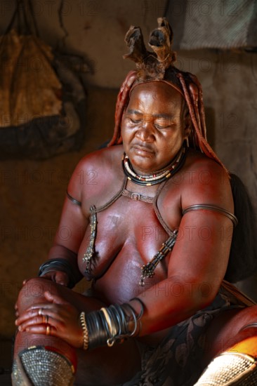 Elderly Himba woman rubbing herself with red paste for skin care, in a hut, traditional Himba