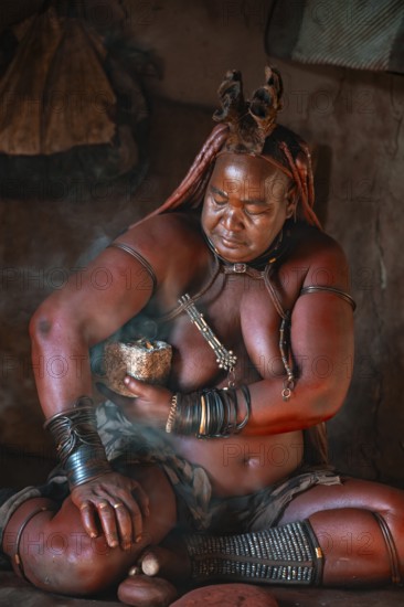 Natural perfume, elderly Himba woman smokes her hair for good smell, in a hut, traditional Himba