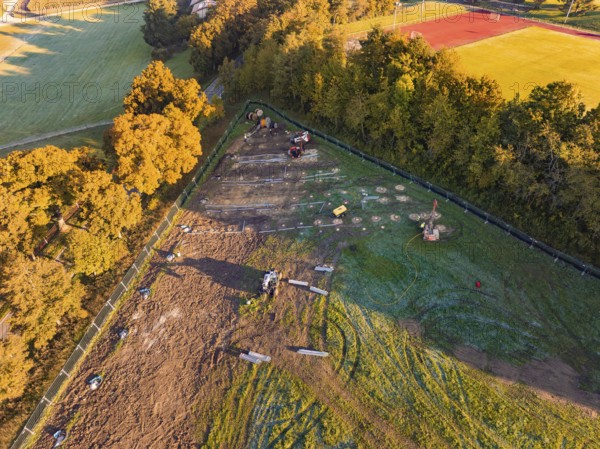 Construction site next to a sports field, surrounded by trees and shade, illuminated by morning