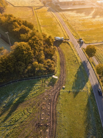 Road through fields with construction, surrounded by trees in early sunlight, construction PV