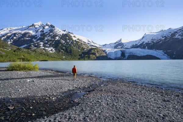 Young man on a pebble beach on a glacial lake, snowy mountains and Portage Glacier glacier on