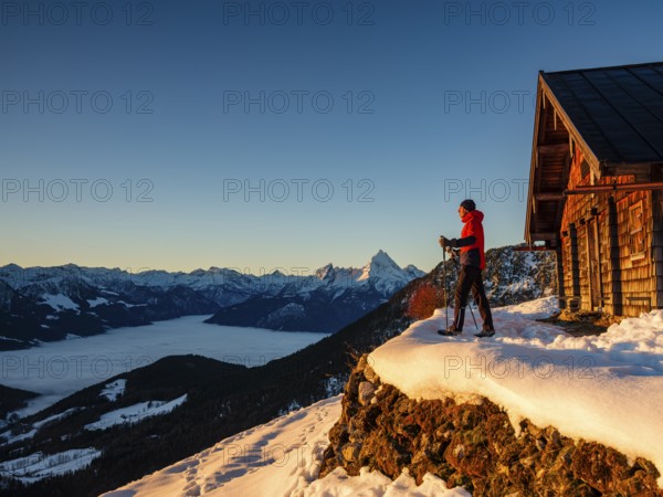 Mountaineers enjoy the sunrise in front of an alpine hut in winter, the Watzmann behind, fog in the