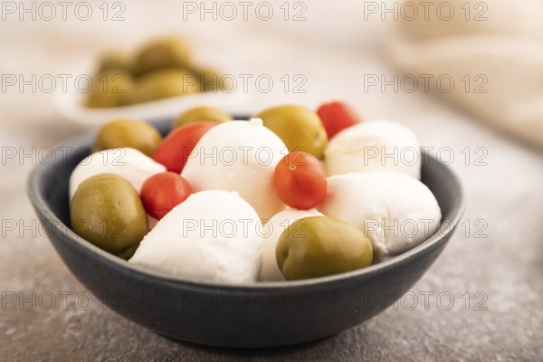 White Mozzarella cheese, with tomatoes and olives in blue bowl on brown concrete background and