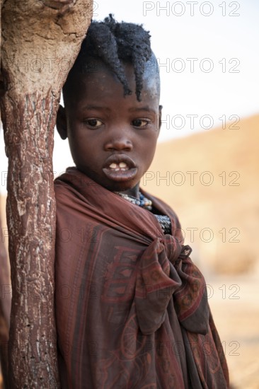 Himba girl leaning at a traditional clay hut, in the morning, traditional Himba village, Kaokoveld,