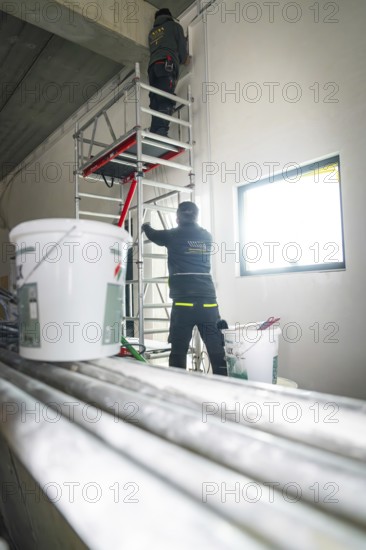 Two workers on scaffolding work on the wall, buckets and pipes are in the foreground, electrician,