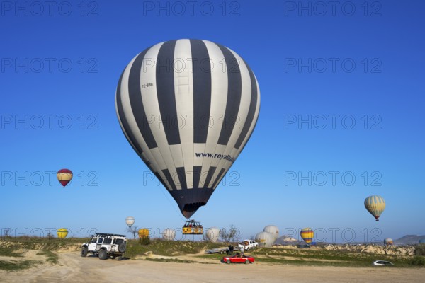 Large striped hot air balloon lands next to jeep and red car in clear landscape, Göreme Tarihî