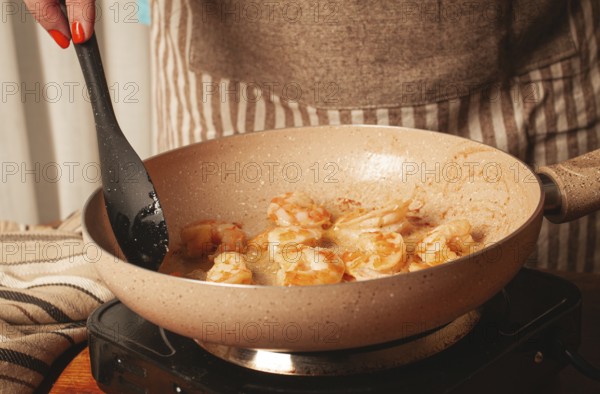 A person is stirring shrimp in a frying pan on the stovetop. The kitchen is warm and inviting, with