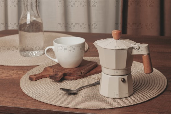 A moka pot sits on a wooden cutting board beside a white cup on a round placemat. A glass bottle of