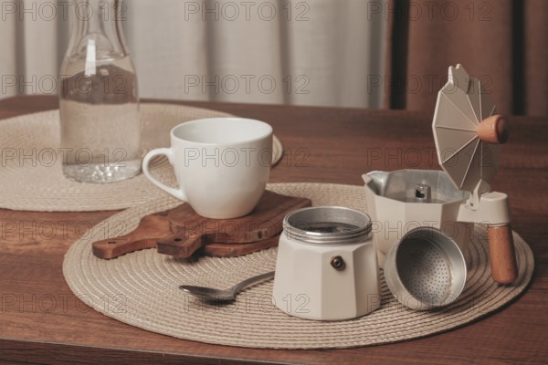 A cozy coffee setup features a white cup, a moka pot, a spoon, and a water bottle on a wooden table