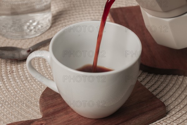 Hot coffee is poured into a white cup resting on a wooden coaster. A serene breakfast setting is