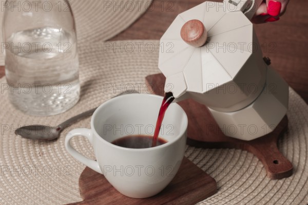 Freshly brewed coffee is being poured from a stovetop espresso maker into a white cup on a wooden