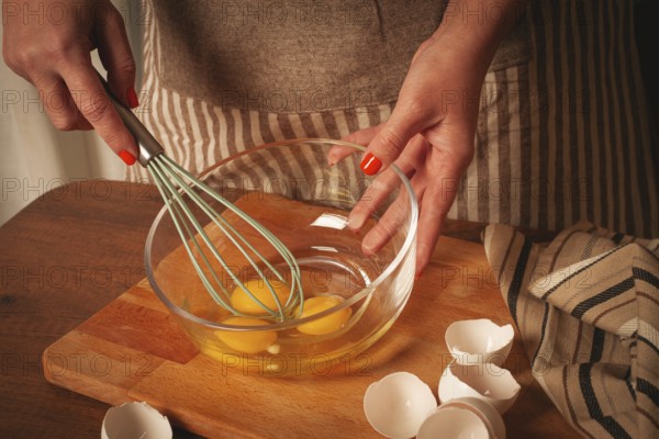 Hands are mixing fresh eggs in a glass bowl with a whisk. Egg shells are scattered on the wooden