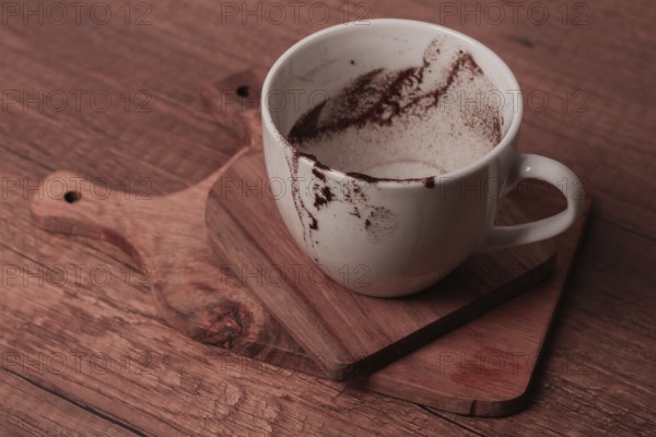 A white cup rests on a wooden cutting board showing remnants of coffee. The cozy setting suggests a