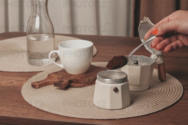 A hand holds a spoonful of coffee grounds over a moka pot while a cup and a bottle of water sit on