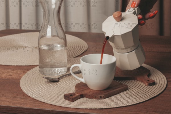 A hand pours freshly brewed coffee from a stovetop maker into a white cup. A glass pitcher filled