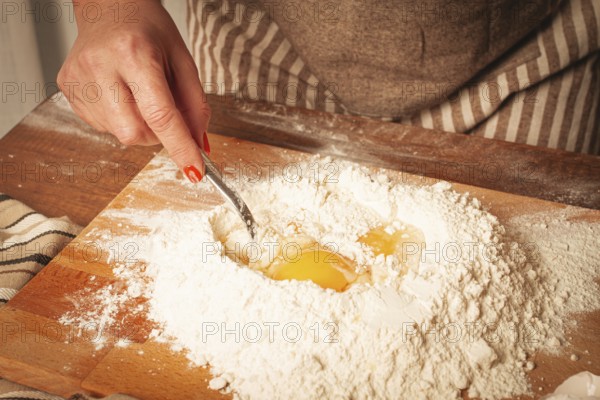 A hand with bright nails mixes eggs into a pile of flour on a wooden board. The cozy kitchen