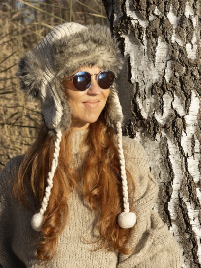 Slender pretty woman with fur hat and pompoms, red hair and warm winter colors between birch forest
