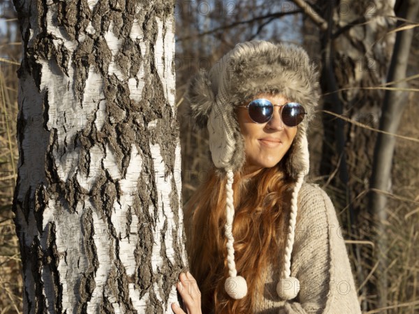 Slender pretty woman with fur hat and pompoms, red hair and warm winter colors between birch forest