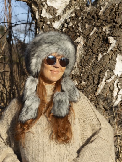 Slender pretty woman with Mongol hat, red hair and warm winter colors between birch forest and