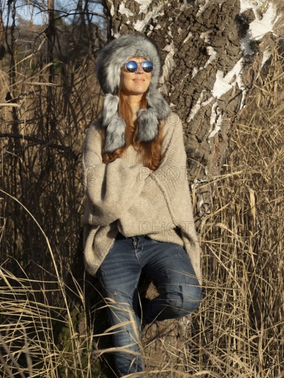 Slim woman with fur hat and sunglasses, red hair and warm winter colors between birch forest and