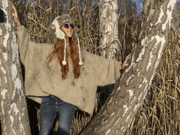 Slender pretty woman with fur hat and sweater, red hair and warm winter colors between birch forest