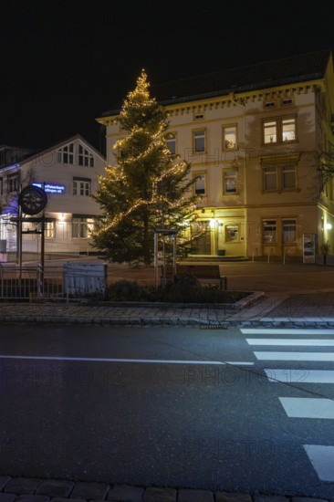 An illuminated Christmas tree in front of a building and a pedestrian crossing at night, Aidlingen,