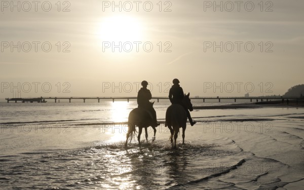 Two female riders ride their horses through the shallow water of the Baltic Sea at sunrise,