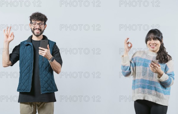 Smiling couple holding a cell phone and gesturing OK, isolated. Happy young couple holding phone